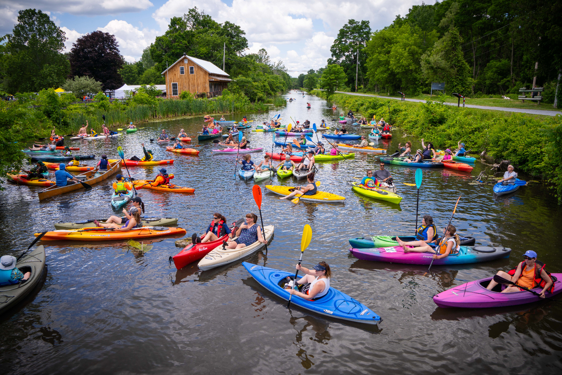 Dozens of kayakers paddling along a river beside a wooded area