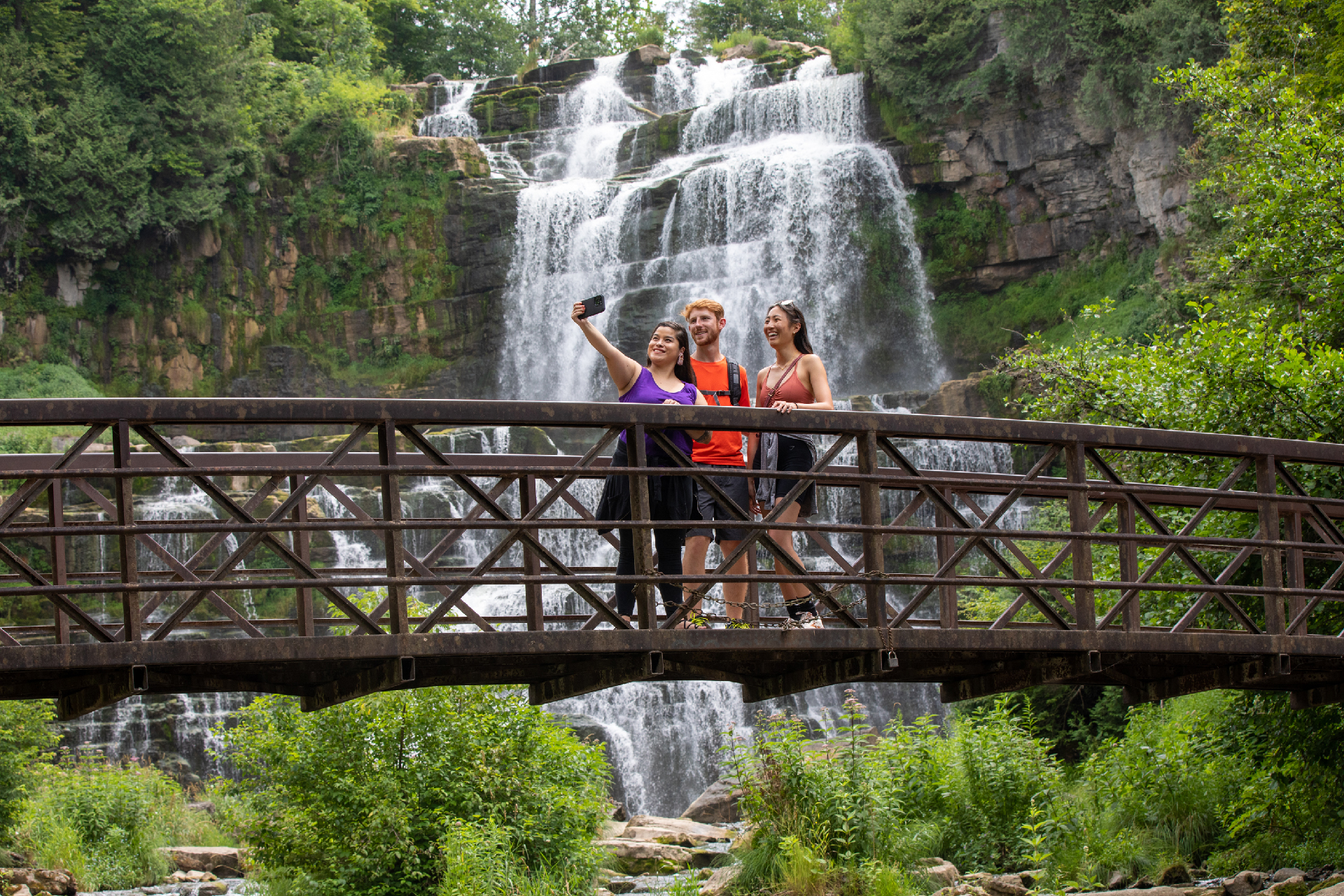 Three hikers taking a selfie while standing on a bridge in front a lush waterfall